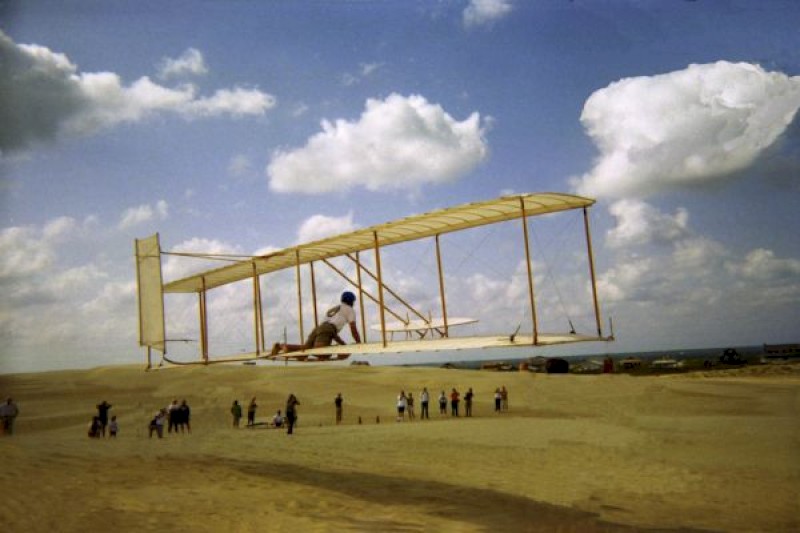 A person is piloting a historic biplane glider over a sandy area with spectators below, under a partly cloudy sky.