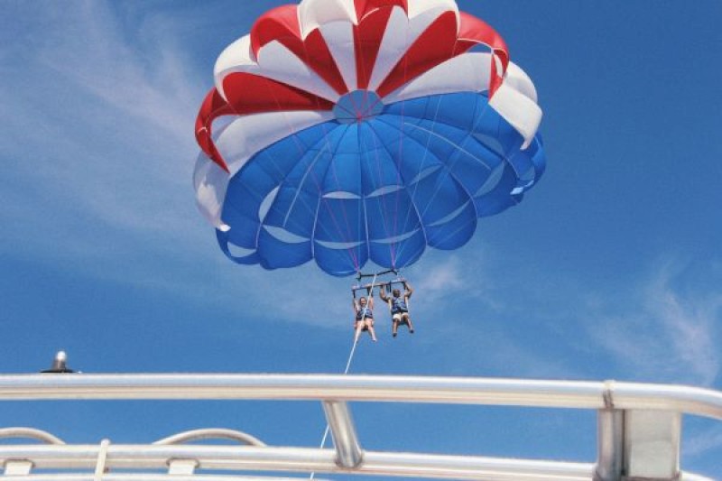 Two people are parasailing above the water with a multicolored parachute. They are tethered to a boat below. The sky is clear and blue.