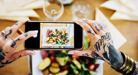 A person with tattooed hands taking a photo of a colorful plate of food using a smartphone, with a table setting and glasses in the background.