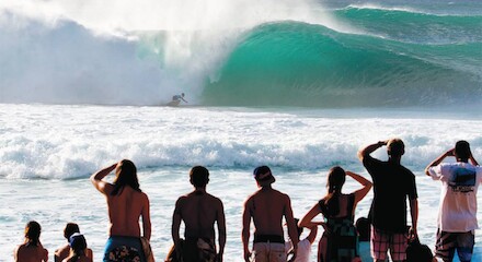 A group of people stand on the beach watching a surfer riding a huge wave.