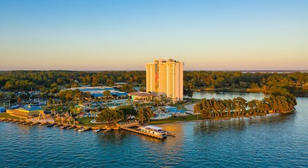 The image shows a tall building complex surrounded by water, trees, and boats, possibly a resort or hotel, with a clear blue sky.