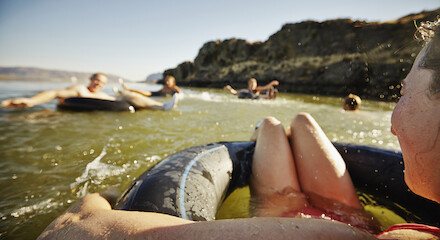 People are floating on inner tubes in a body of water, surrounded by rocky terrain and sunny skies.