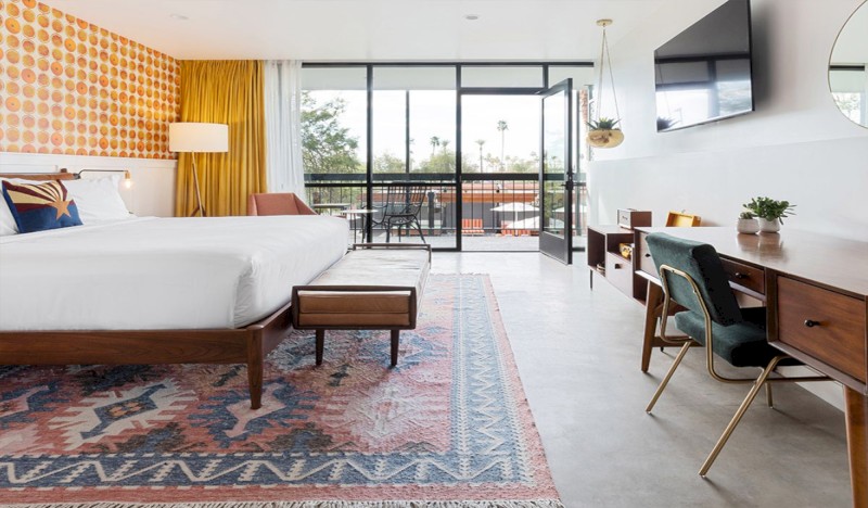 Una habitación de hotel moderna cuenta con una cama grande, una alfombra estampada, un escritorio, sillas y un televisor montado en la pared, con un balcón que ofrece vistas al exterior.