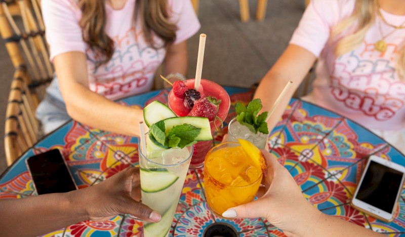 Cuatro personas brindando con bebidas coloridas en una mesa vibrante, cada una sosteniendo un vaso único. Sus teléfonos están colocados en el borde de la mesa.