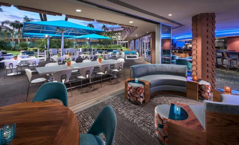 The image shows a modern outdoor dining area with blue umbrellas and a nearby indoor lounge with stylish seating and ambient lighting.