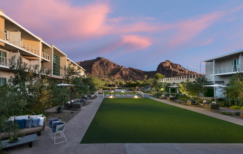 The image showcases a landscaped courtyard between two buildings, with seating areas, greenery, and a view of mountains under a pinkish sky.