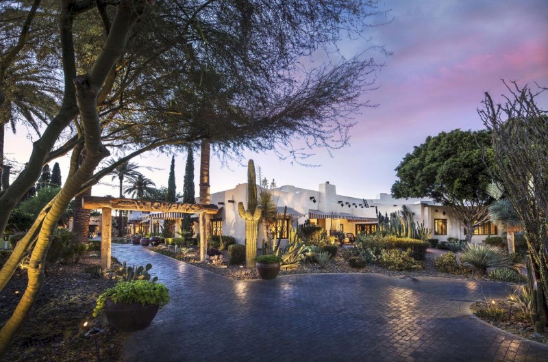 The image shows a beautifully landscaped desert resort at dusk, with cacti, trees, a pathway, and a white adobe-style building illuminated by soft lights.