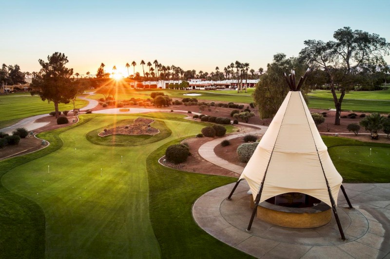 The image shows a landscaped golf course at sunset with a teepee structure and winding paths, surrounded by trees and manicured lawns.