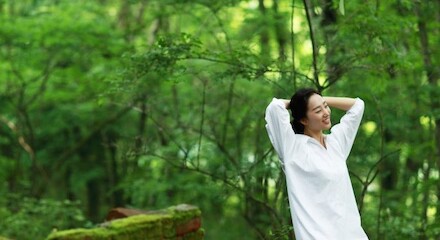 A woman in a white shirt stands in a lush forest, smiling and stretching with her hands behind her head, enjoying the natural surroundings.