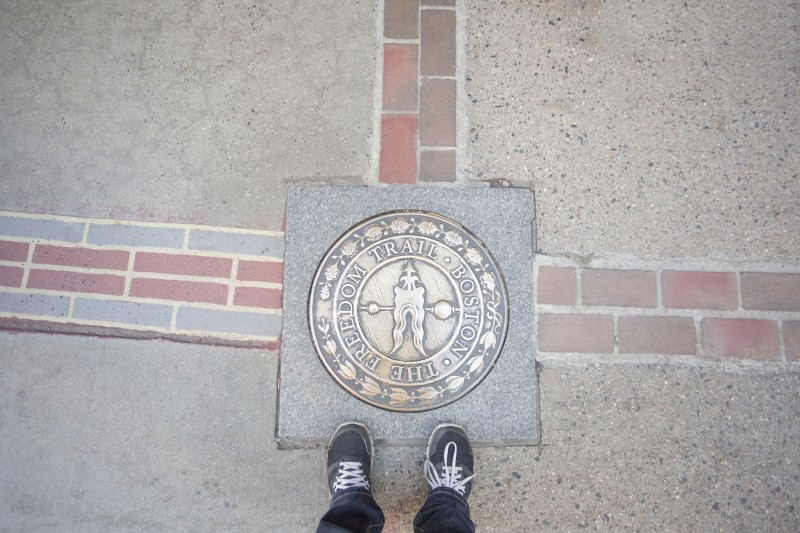 Feet standing on a marker for "The Freedom Trail, Boston," embedded in the sidewalk, bordered by a line of bricks.
