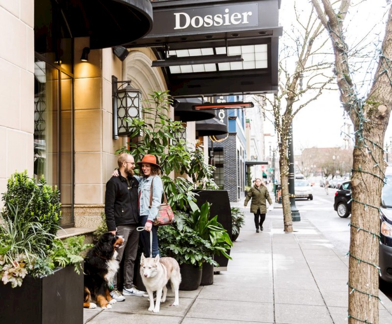 A couple with two dogs stands outside a building with a "Dossier" sign. The street is lined with plants and other pedestrians.