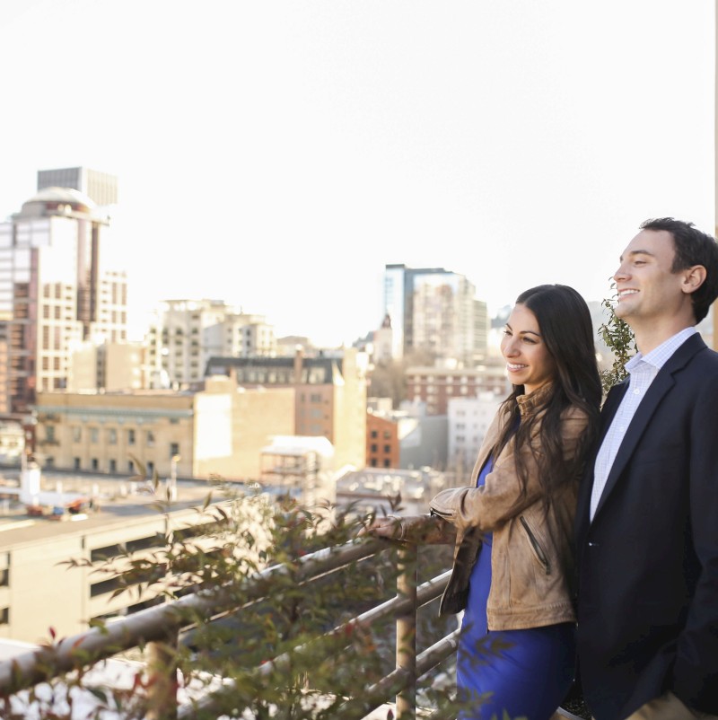 A man and woman are standing on a balcony, smiling, with an urban cityscape in the background. They appear to be enjoying the view together.