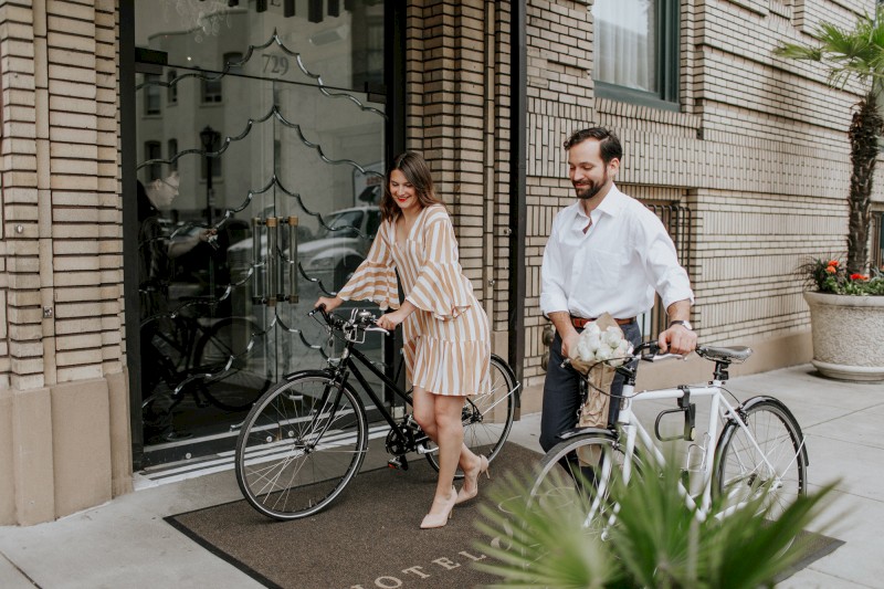 A woman and a man are walking their bicycles out of the hotel entrance onto the sidewalk near a brick building, with plants nearby.