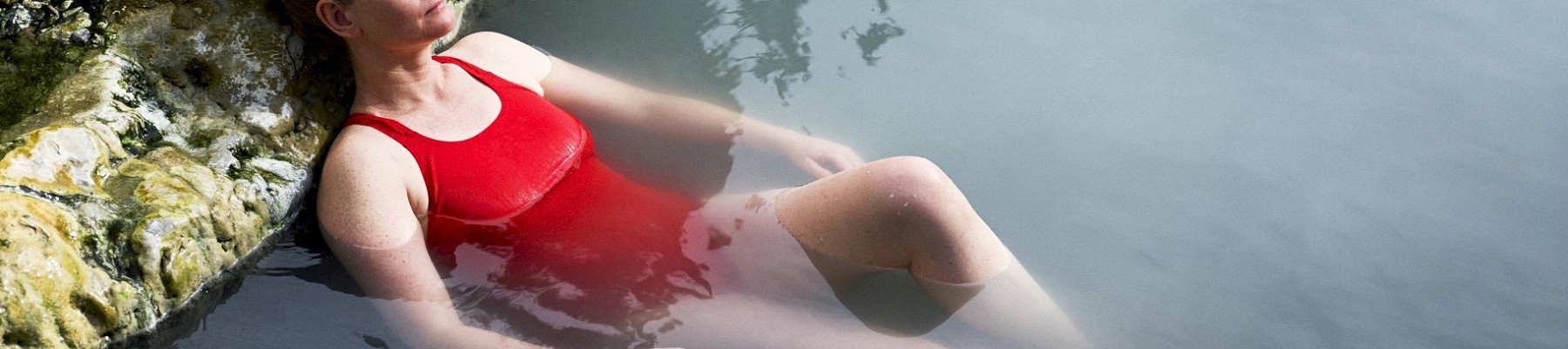 A person in a red swimsuit is floating and relaxing in a natural hot spring, with rocky surroundings in the background.