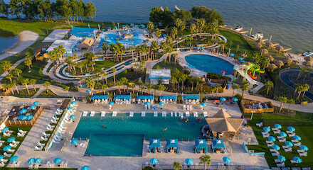Aerial view of a sprawling resort featuring multiple swimming pools, lounge areas with umbrellas and chairs, and a lakeside setting with lush greenery.