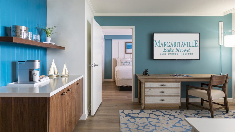 The image depicts a hotel room at Margaritaville Lake Resort. It features a desk, a bed, a coffee maker, and a sign indicating the resort's location.