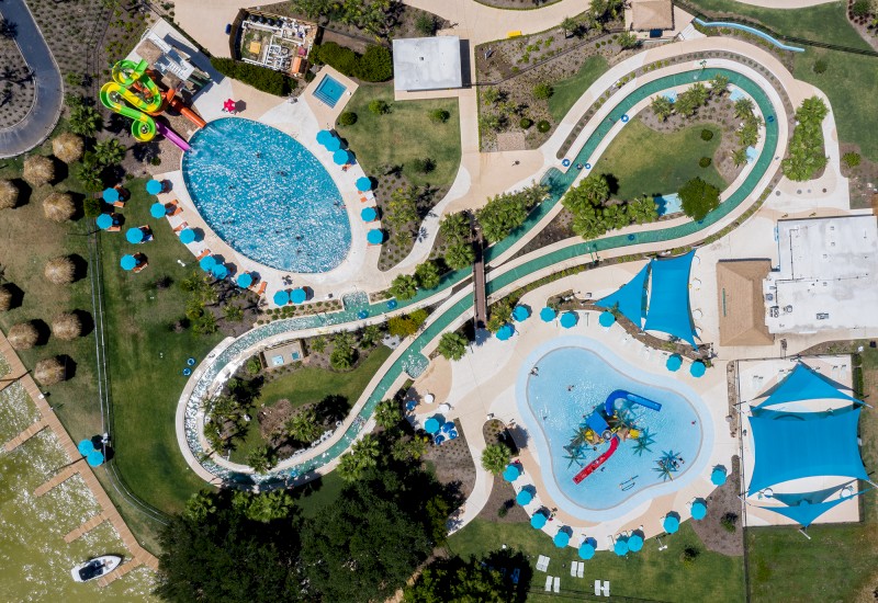 This aerial image shows an outdoor water park with multiple pools, water slides, and a lazy river, surrounded by green spaces and seating areas.