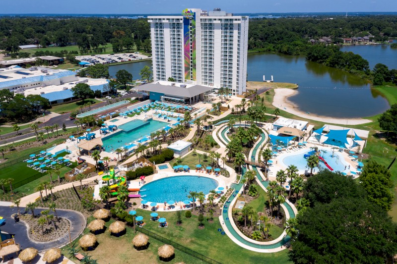 Aerial view of a large resort with multiple pools, water slides, loungers, a lake, and a high-rise building in a lush green landscape.