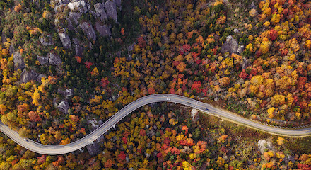 A winding road cuts through a dense forest with vibrant autumn foliage and rocky outcrops. The trees show various shades of orange, yellow, and red hues.