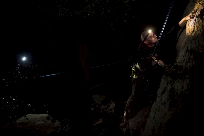 A person wearing a headlamp is climbing a rock or cliff in a dark environment, while another light source is visible in the background.