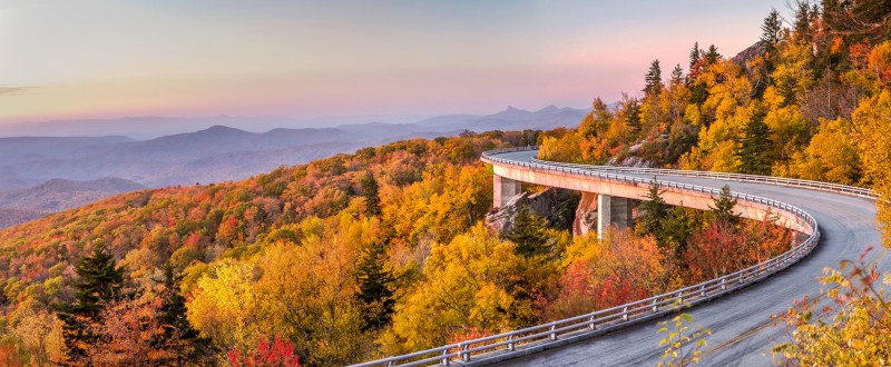 A curving mountain road surrounded by vibrant autumn foliage, with distant blue ridges under a colorful sunset sky.