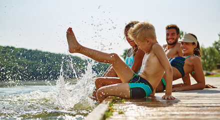 A group of people, including a child splashing water, are sitting on a wooden dock by a body of water, enjoying a sunny day outdoors.