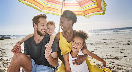 A happy family sits on the beach under a colorful umbrella, smiling and enjoying their time together by the sea.