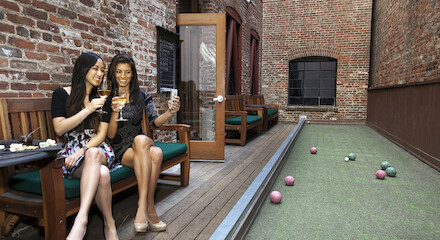 Two people are enjoying drinks on an outdoor patio next to a bocce ball court in an urban setting with brick walls and wooden furniture.