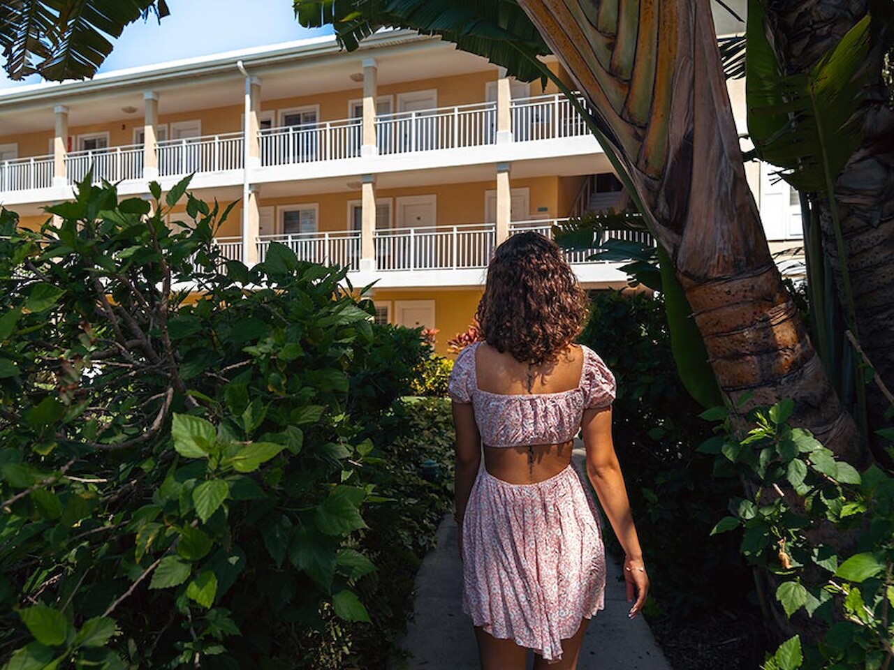 A person in a summer dress walks on a path surrounded by lush greenery, approaching a multi-story building with balconies and railings.