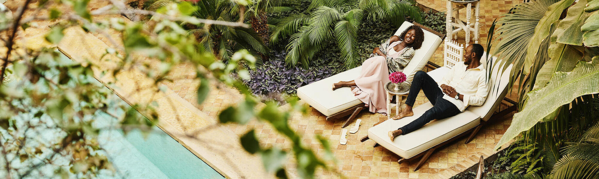 Dos personas están descansando en las sillas junto a la piscina rodeadas de exuberantes plantas tropicales, disfrutando de un momento de relax.