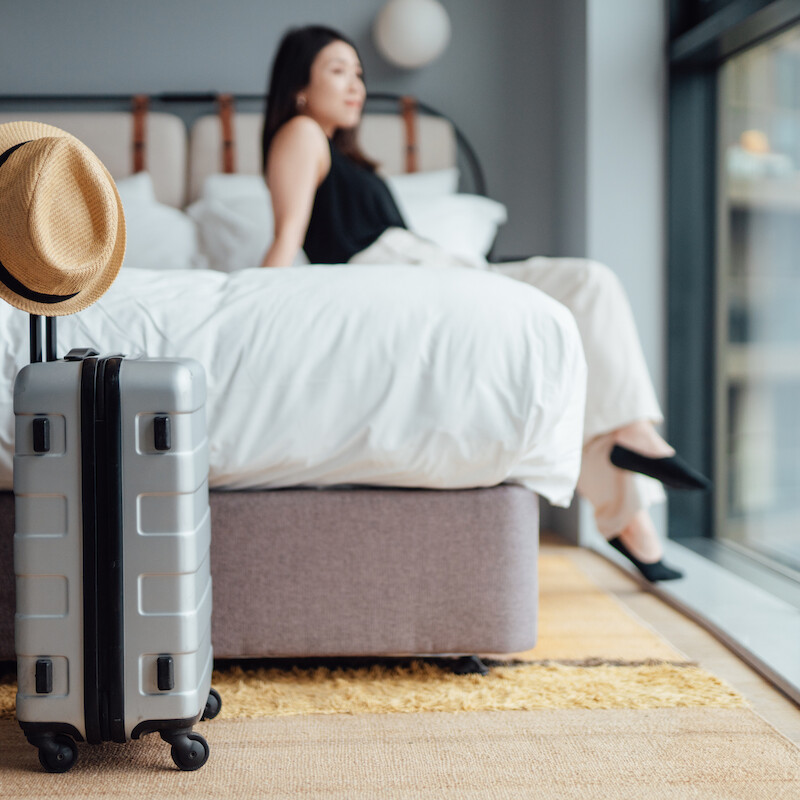 Una mujer sentada en una cama junto a un gran ventanal, con una maleta gris y un sombrero en primer plano, sugiriendo que está de viaje o que ha llegado.