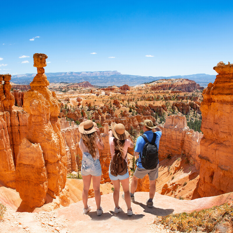 Tres personas con mochilas observan, admirando las formaciones rocosas rojas y los Hoodoos de un paisaje desértico bajo un cielo azul claro.