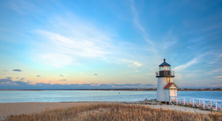 A serene coastal scene with a small lighthouse by the water's edge, surrounded by dry grasses under a clear, pastel sky during sunset.