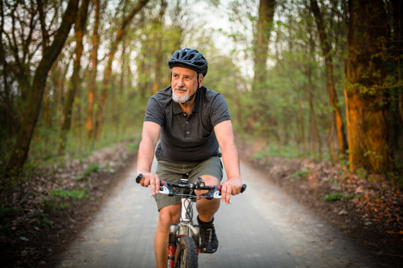 A man wearing a helmet and casual clothes riding a bicycle on a wooded path, surrounded by trees and greenery.