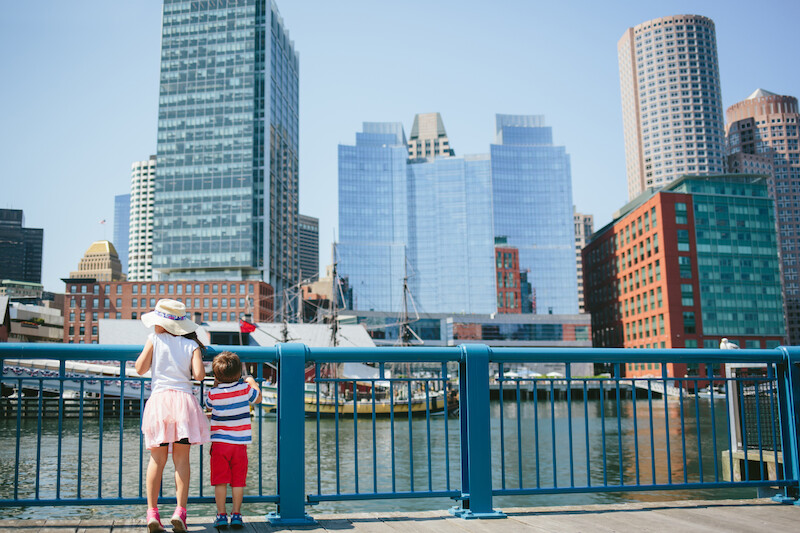 The image shows a woman and a child standing by a blue railing, overlooking water with boats and a cityscape of tall buildings in the background.