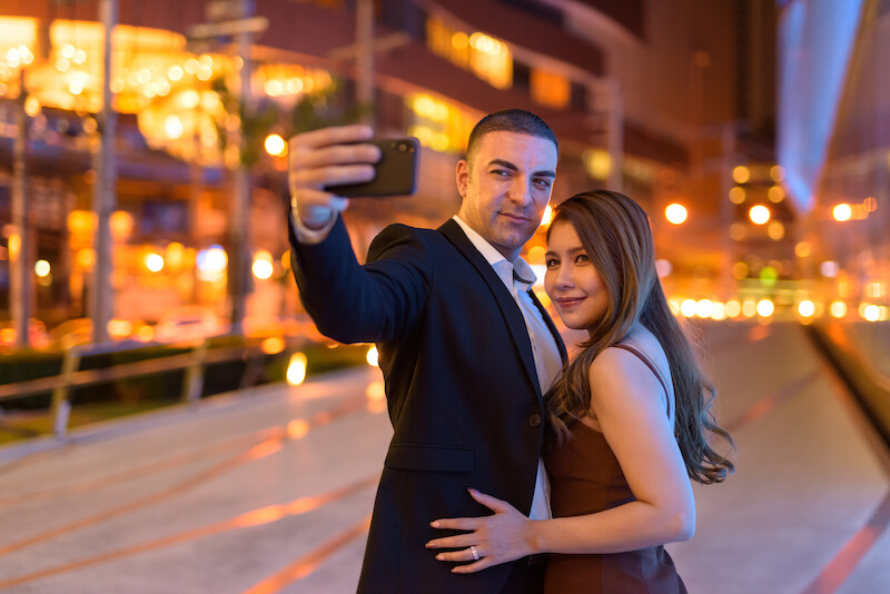 A couple is taking a selfie at night in an urban area with bright city lights in the background.