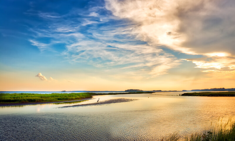 The image depicts a serene landscape with a calm body of water, a thin strip of land, green vegetation, and a vibrant sky with clouds during sunset.