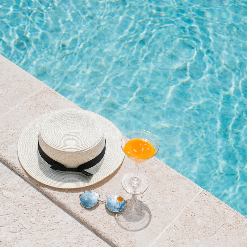 A straw hat, sunglasses, and a glass of orange cocktail sit beside a clear, blue swimming pool on a sunny day.