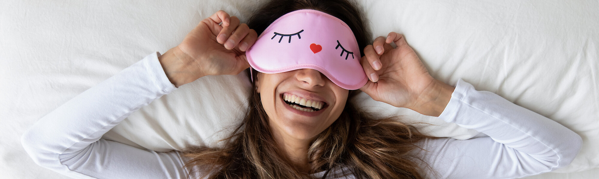A woman is lying in bed wearing a white shirt and a pink sleep mask, smiling broadly while stretching and holding the mask over her eyes.