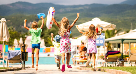 Four children joyfully run towards a swimming pool, holding flotation devices. Mountains and poolside lounging chairs are in the background, under sunny skies.