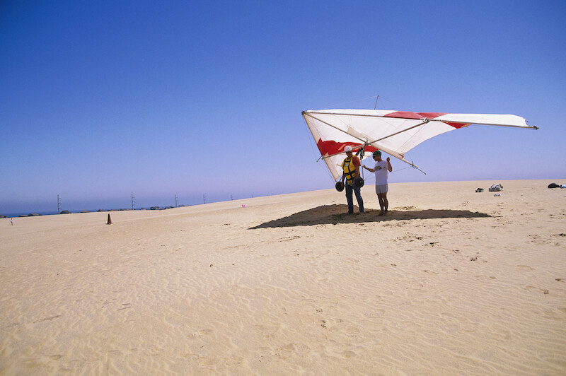 Two individuals are ready for hang gliding on a sandy beach under a clear blue sky, holding the glider's frame in preparation for takeoff.