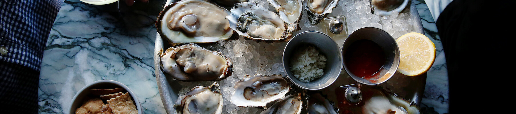 A platter of oysters on a table, accompanied by beverages, crackers, lemon, and sauce, with hands holding glasses in the background.