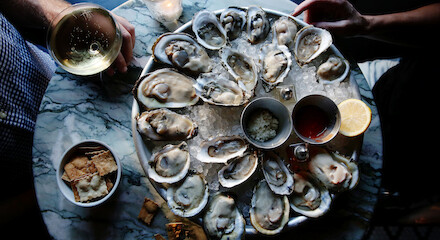 A platter of oysters on a table, accompanied by beverages, crackers, lemon, and sauce, with hands holding glasses in the background.