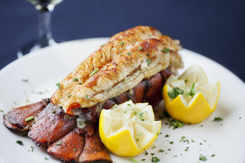 This image shows a cooked lobster tail garnished with herbs, served with two lemon wedges on a white plate, with a glass of wine in the background.
