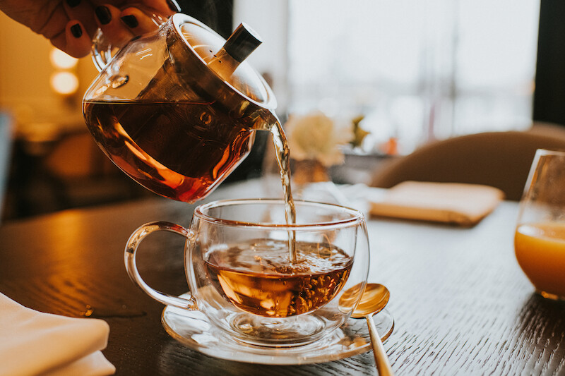 A hand is pouring tea from a glass teapot into a clear teacup on a table. A spoon and a napkin are nearby, and the background is slightly blurred.