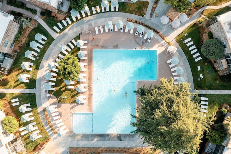 Aerial view of a rectangular swimming pool surrounded by lounge chairs and greenery in a well-organized layout. Trees provide shade nearby.