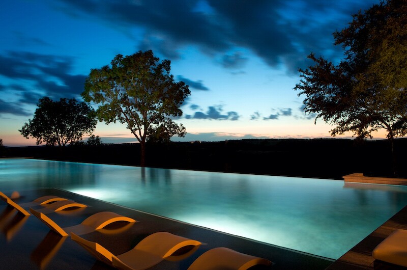 The image shows an outdoor pool area at dusk with lounge chairs aligned by the pool, trees in the background, and a serene sky.