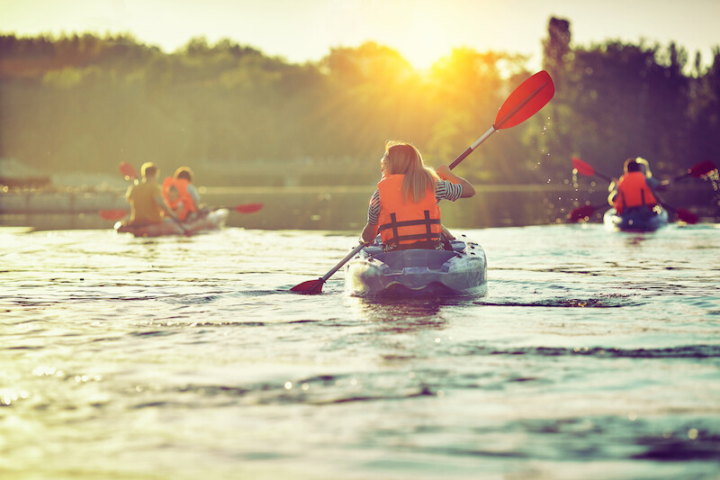 Several people are kayaking on a lake, with the sun setting in the background. They wear life vests and enjoy the serene evening water.