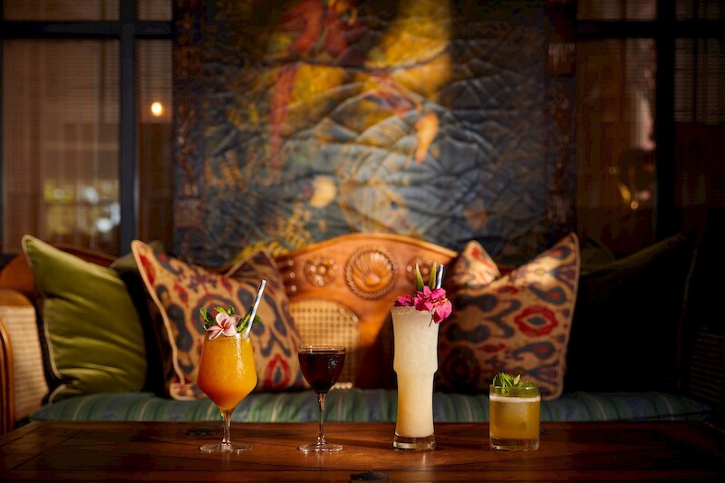 The image shows a cozy setting with decorative pillows on a couch and four colorful drinks on a table in front of it.