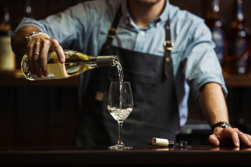 A person wearing a blue shirt and apron is pouring white wine into a glass on a bar counter, with a cork and bottle opener beside them.